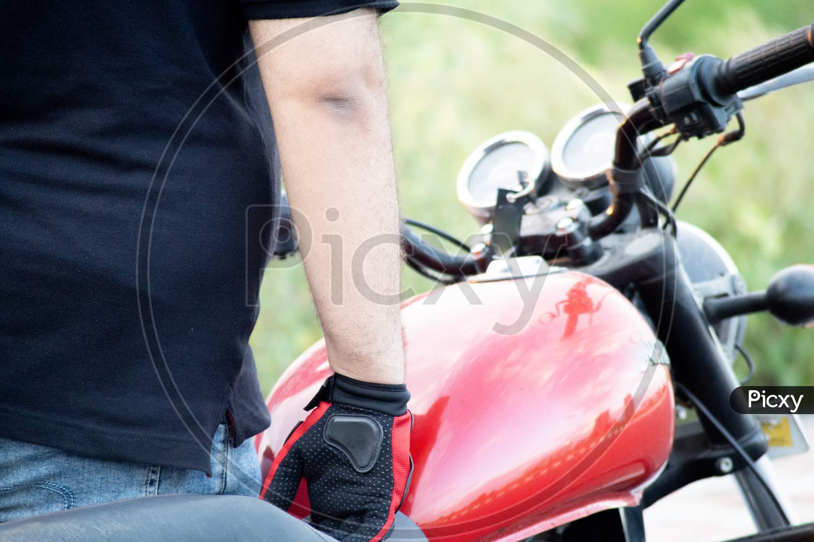 Young Indian Man Wearing Red Riding Gloves With Protection While Holding The Handle Bars Of A Motorcycle With A Red Tank