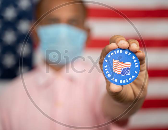 Man In Medical Mask Showing I Voted By Mail Sticker With Us Flag As Background - Concept Of Mail In Voting At Usa Election.