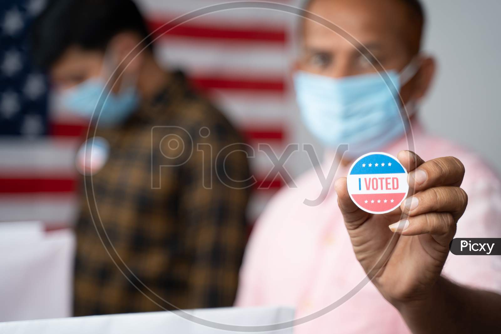 Close Up Of Hands, Man In Medical Mask Showing I Voted Sticker At Polling Booth With Us Flag As Background - Concept In Person Voting At Us Election.