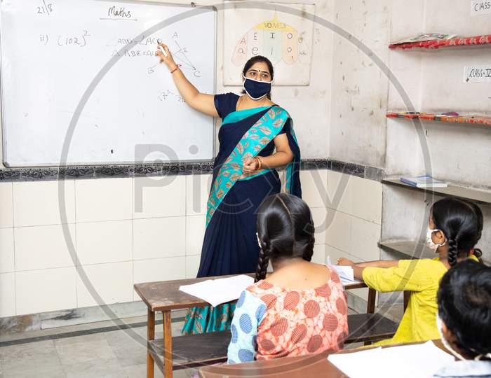 Indian Teacher And Students Wearing Face Masks Maintaining Social Distancing Study In Classroom Back At School During Covid19 Pandemic.