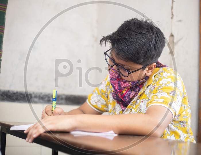 Indian Boy Student Studying Writing In Classroom Wearing Mask Maintaining Social Distancing Looks At Camera, School Reopen During Covid19 Pandemic