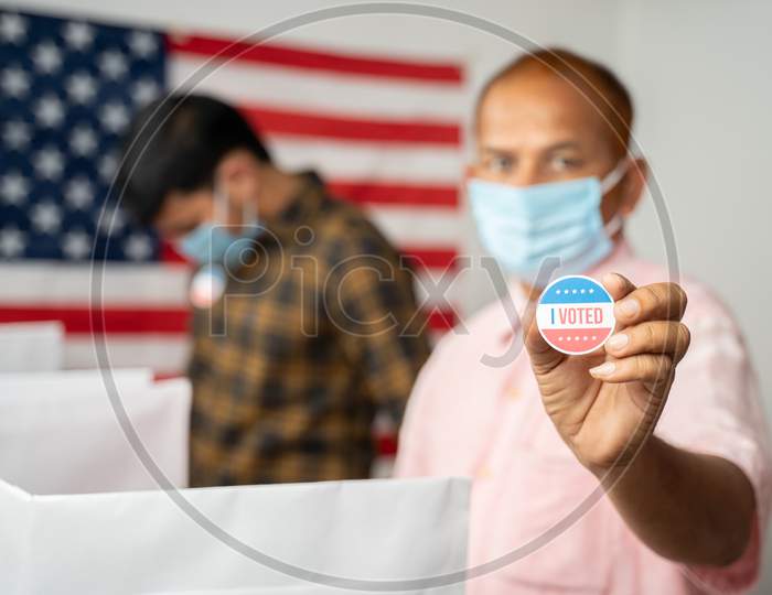 Man In Medical Mask Showing I Voted Sticker At Polling Booth With Us Flag As Background - Concept In Person Voting At Us Election.