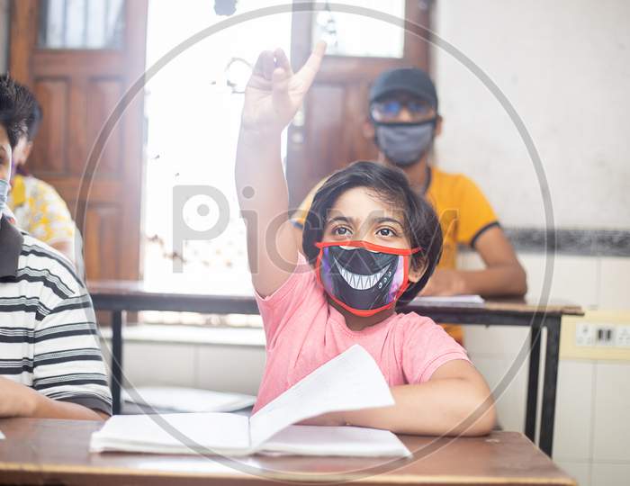 Indian Students Raise Hand Studying In Classroom Wearing Mask Maintaining Social Distancing Looks At Camera, School Reopen During Covid19 Pandemic, New Normal. Selective Focus