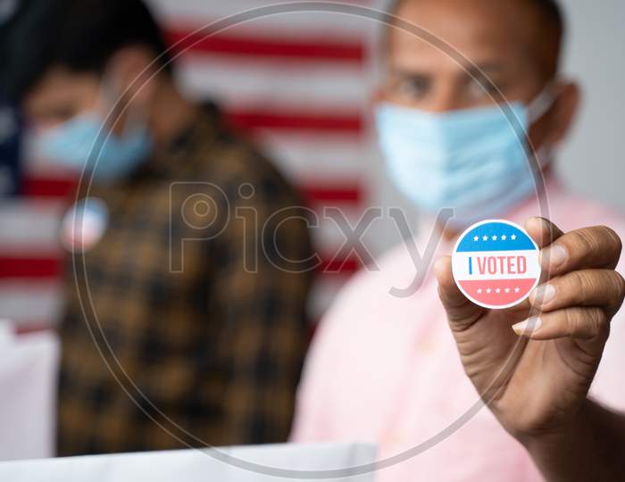 Close Up Of Hands, Man In Medical Mask Showing I Voted Sticker At Polling Booth With Us Flag As Background - Concept In Person Voting At Us Election.