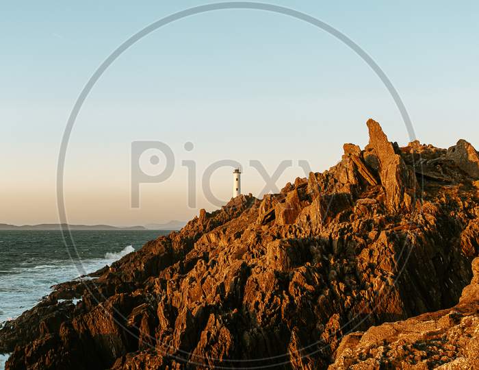 White Lighthouse Over Huge Risky Rocks In The Coastline Of Spain During A Sunny Day Of Autumn