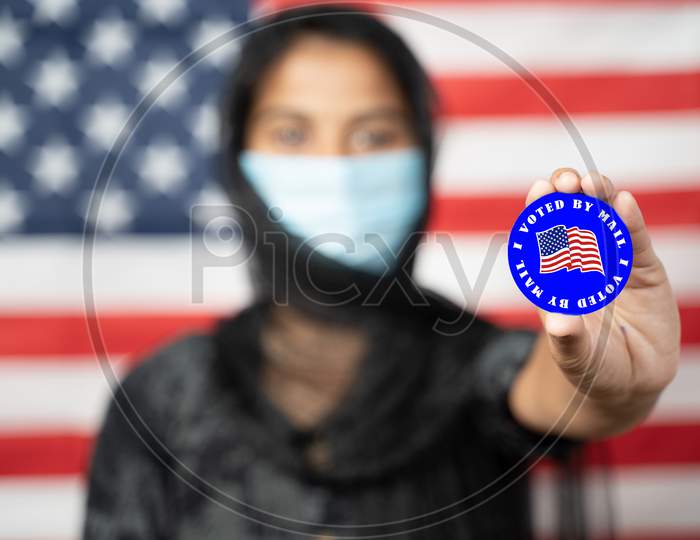 Selective Focus On Hands, Girl With Hijab Or Head Covering And Mask Worn Showing I Voted Sticker With Us Flag As Background - Concept Of Voting During Us Election.