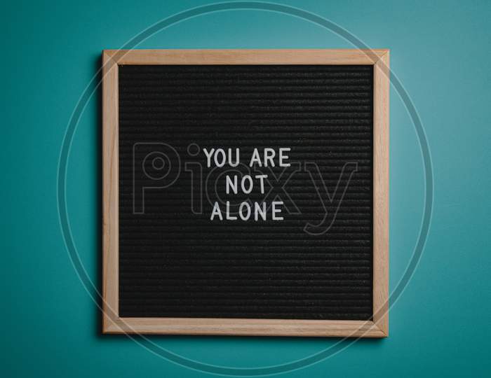 Life's Quote " You Are Not Alone "  In Black Board on Brown Wooden Frame Behind Green Background.