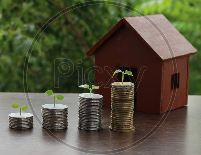 Model Of House With Money Coins Stack On Wooden Table On Blurred Background. Growing Money - Plant On Coins - Saving And Investment Concept.