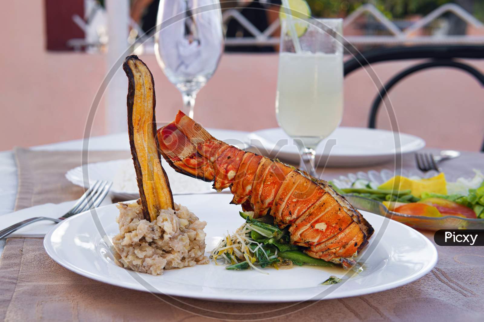Grilled Lobster Is Beautifully Placed On A Plate Along With A Side Dish Of Mashed Potatoes, Fried Banana, Vegetables, Rice And A Glass Of Cocktail. Expensive Dinner At A Restaurant