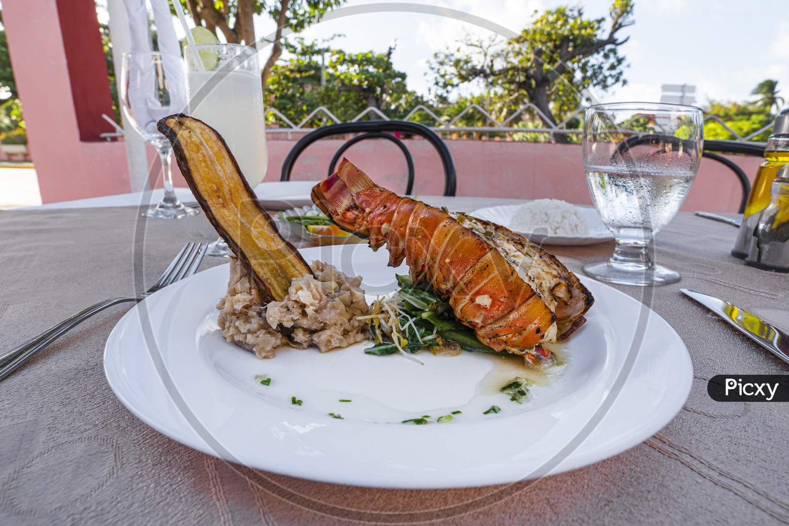 Grilled Lobster Is Beautifully Placed On A Plate Along With A Side Dish Of Mashed Potatoes, Fried Banana, Vegetables, Rice And A Glass Of Cocktail. Expensive Dinner At A Restaurant Outdoor