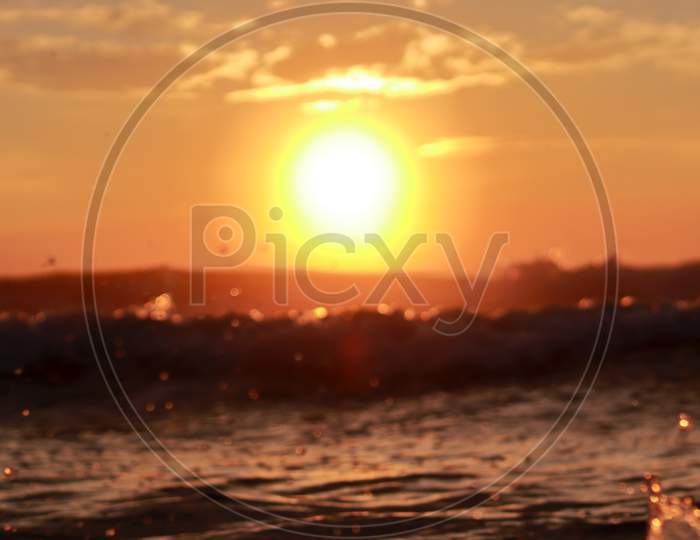 A Defocused Background In Which A Which Bright Round Sun Rises Above The Sky With The Rising Waves Of The Sea