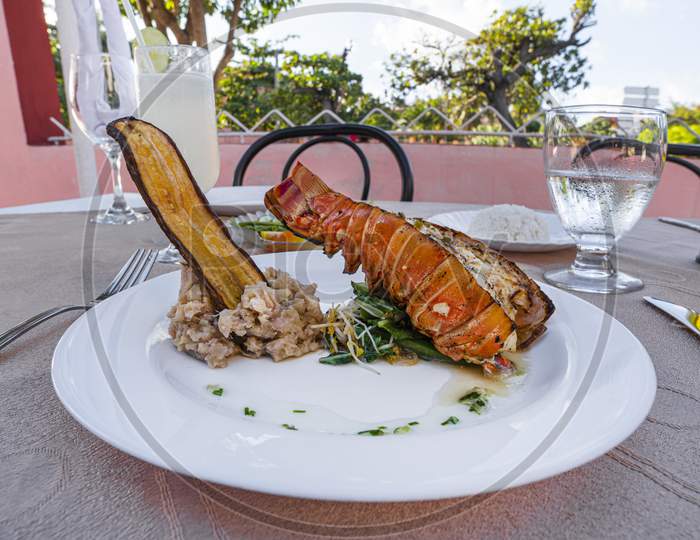 Grilled Lobster Is Beautifully Placed On A Plate Along With A Side Dish Of Mashed Potatoes, Fried Banana, Vegetables, Rice And A Glass Of Cocktail. Expensive Dinner At A Restaurant Outdoor