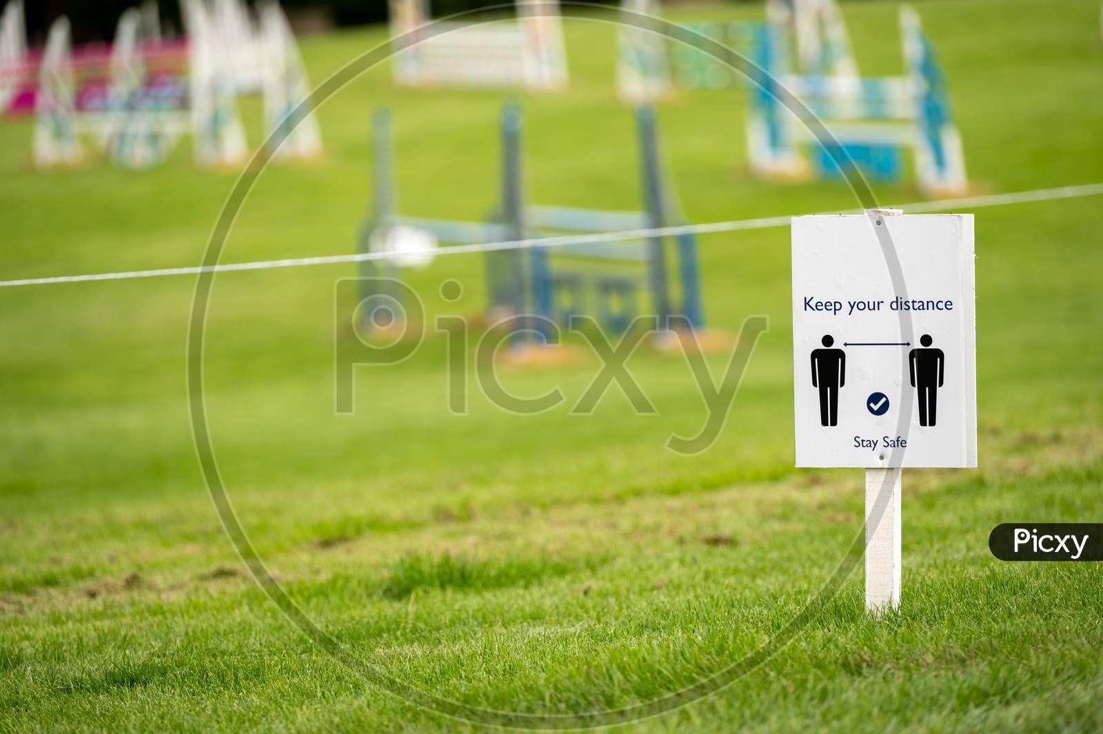 A Covid 19 Social Distancing Sign Staked Into Grass With An Horse Jumps At An Equestrian Show Jumping Outdoor Event In The Background