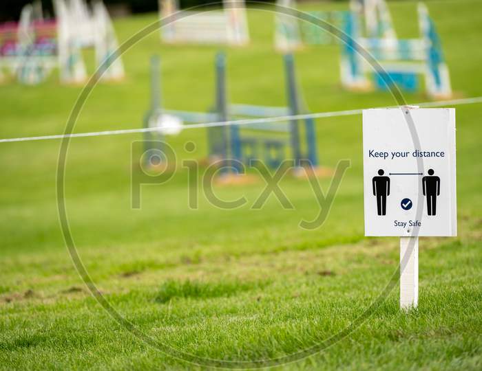 A Covid 19 Social Distancing Sign Staked Into Grass With An Horse Jumps At An Equestrian Show Jumping Outdoor Event In The Background