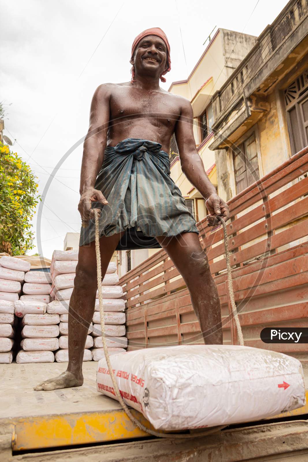 Indian Construction Worker Looking At Camera And Smiling Before Lifting A Sack Of Cement