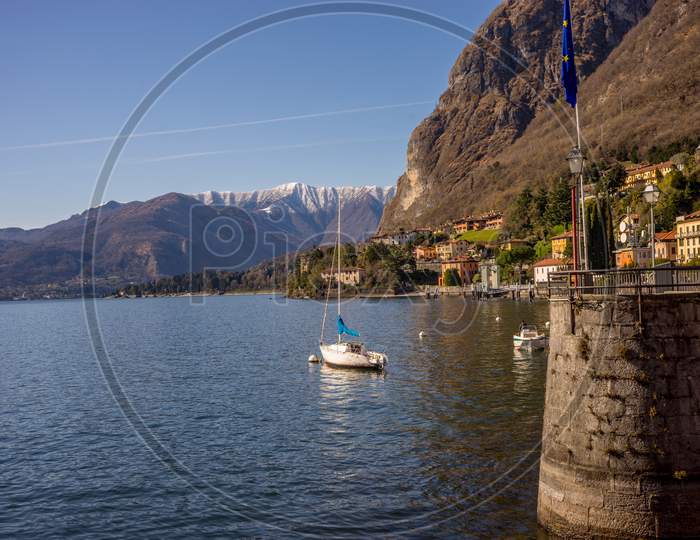 Italy, Menaggio, Lake Como, A Small Boat In A Body Of Water With A Mountain In The Background