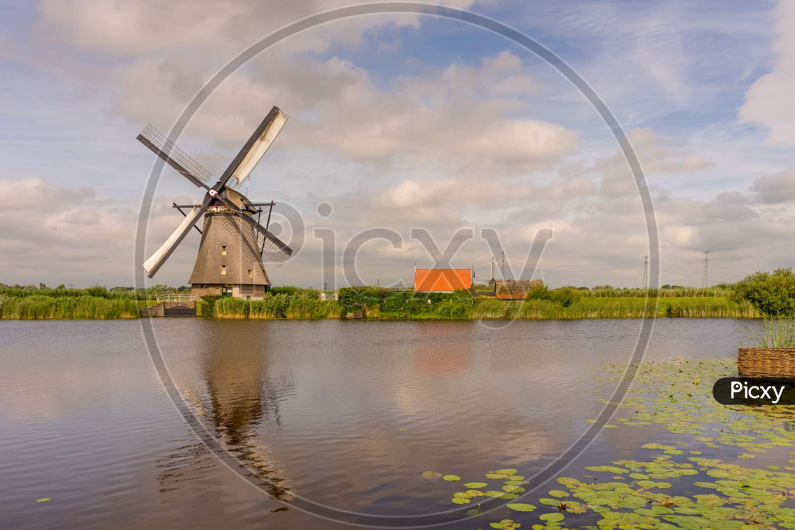 Netherlands, Rotterdam, Kinderdijk, Heritage Windmill Above Lush Green Grass Along A Canal With Reflection