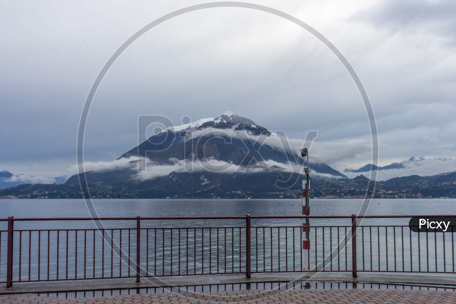 Italy, Varenna, Lake Como, A Body Of Water With A Snowcap Mountain In The Background