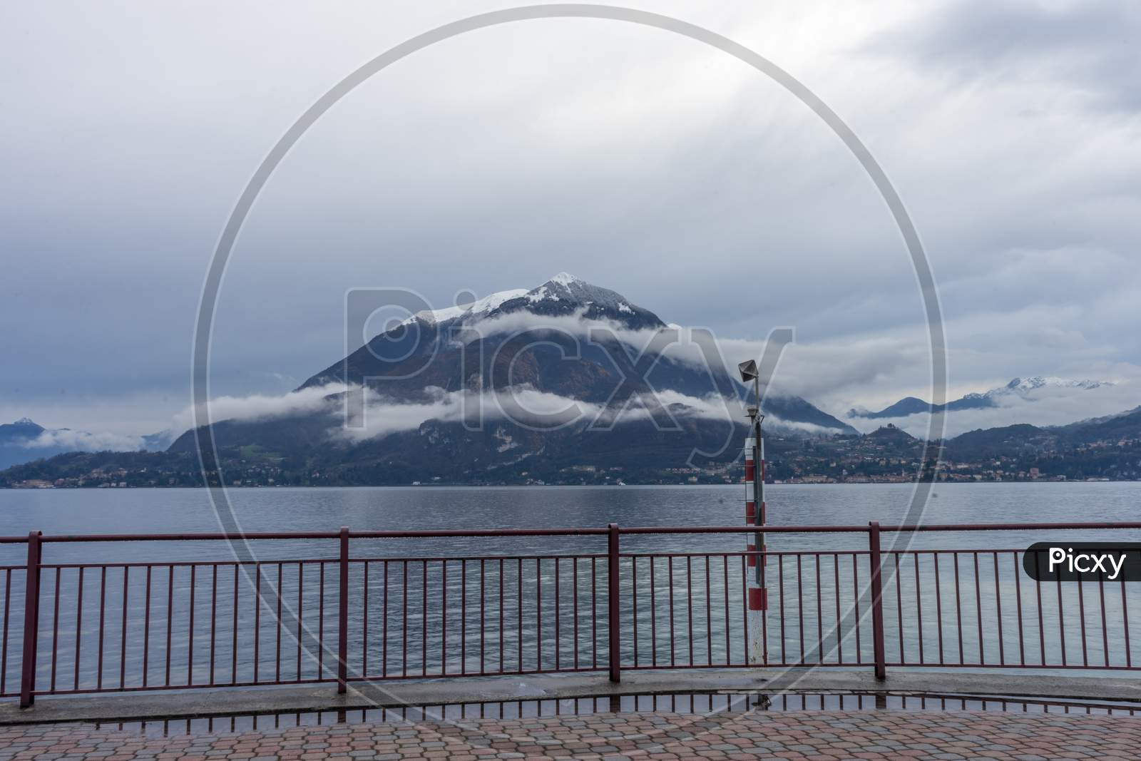 Italy, Varenna, Lake Como, A Body Of Water With A Snowcap Mountain In Front Of A Fence