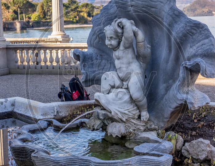 Italy, Bellagio, Lake Como, Abellagio, Italy-April 1, 2018: Water And Lily Fountain At Giardini Di Villa Melzi Group Of Stuffed Animals Sitting Next To A Body Of Water