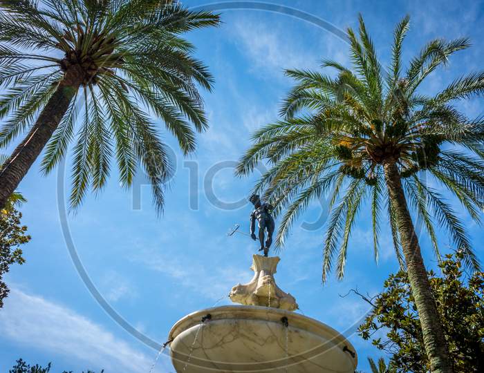 Neptune Under The Palm Tree In Alcazar Garden With A Blue Sky In Seville,  Spain, Europe