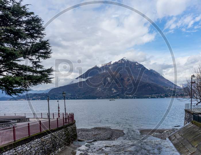 Italy, Varenna, Lake Como, A Body Of Water With A Mountain In The Snow