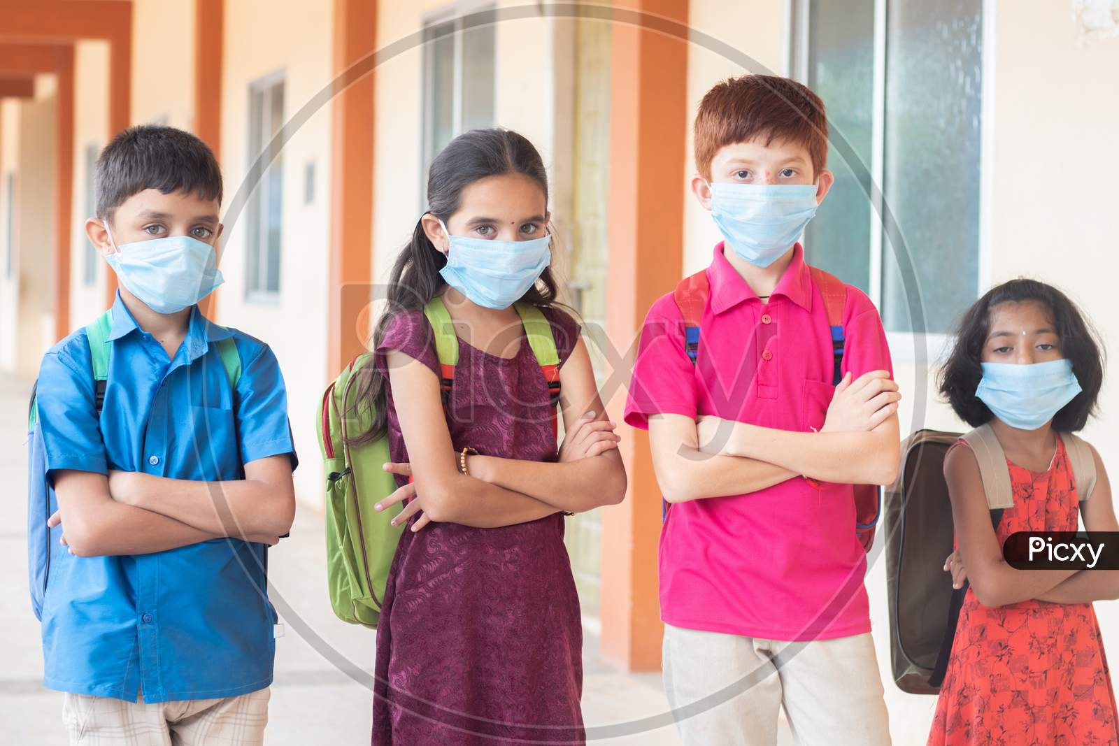 Children In Medical Mask With School Backpack Standing With Arms Crossed By Looking To The Camera- Concept Of Back To School And Reopen.