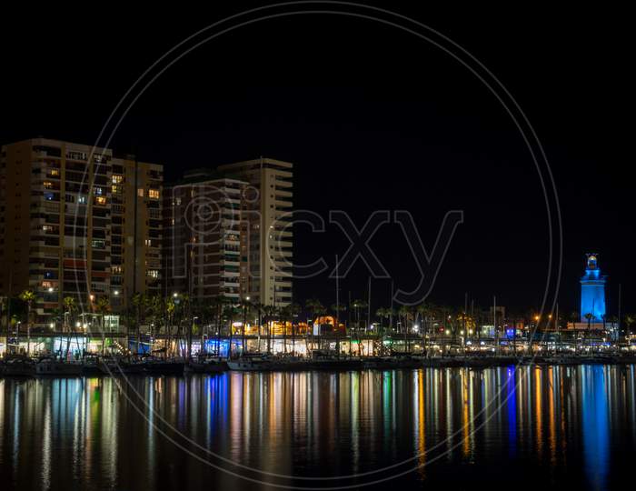 View Of Malaga City And Lighthouse And Their Reflections On Water From Harbour, Malaga, Spain, Europe At Night