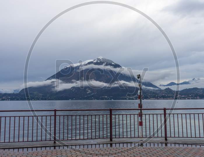Italy, Varenna, Lake Como, A Body Of Water With A Snowcap Mountain In Front Of A Fence