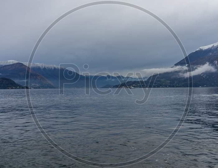 Italy, Varenna, Lake Como, A Body Of Water With A Mountain In The Background