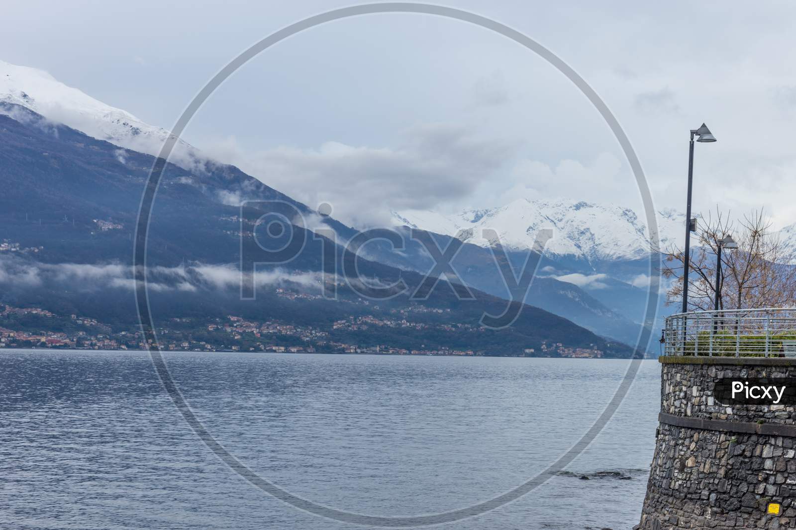 Italy, Varenna, Lake Como, A Large Body Of Water With A Snowcap Mountain In The Background