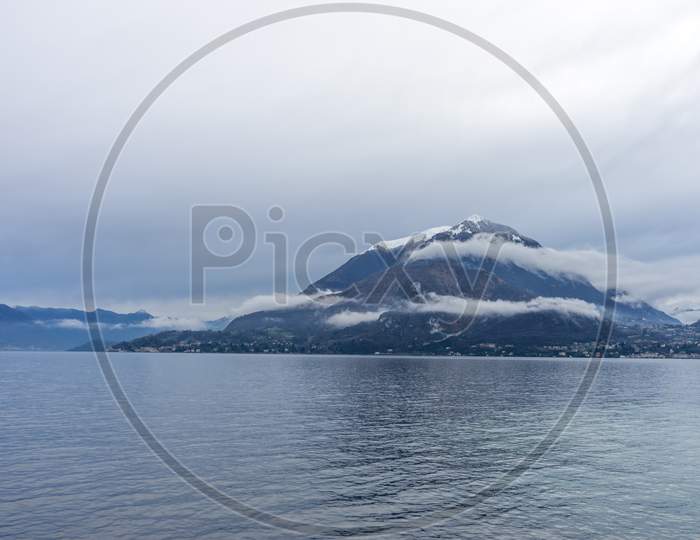 Italy, Varenna, Lake Como, A Large Body Of Water With A Snowcap Mountain In The Background