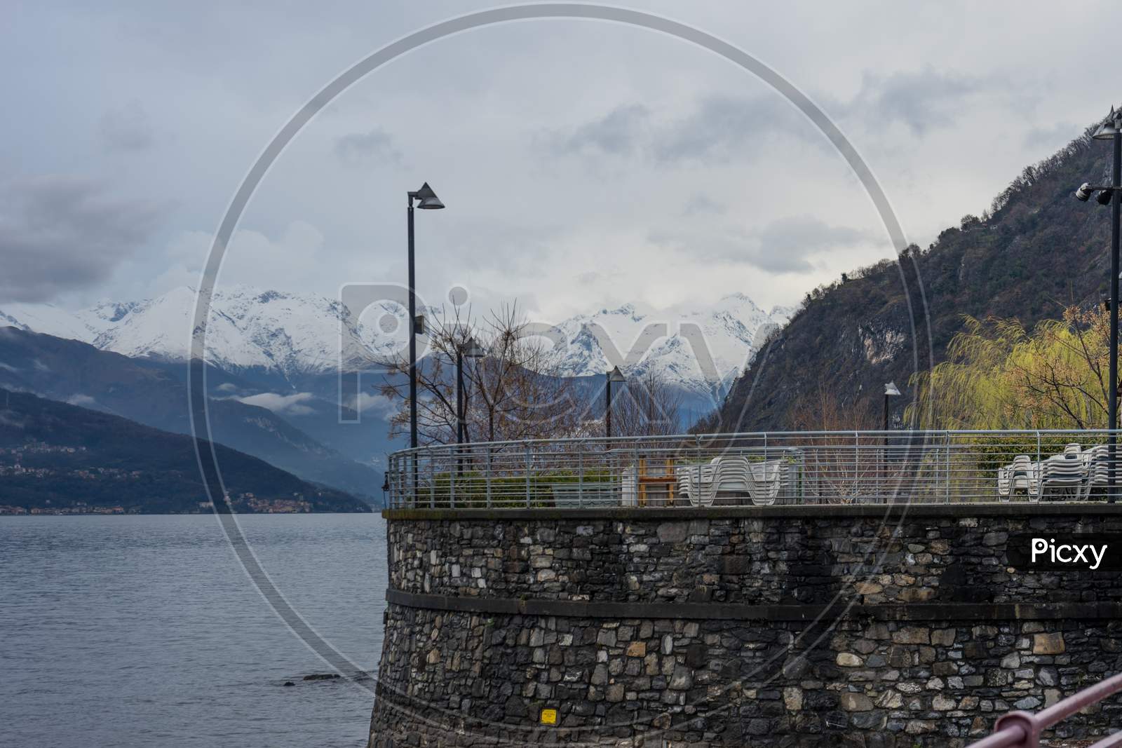 Italy, Varenna, Lake Como, A Bridge Over A Body Of Water With A Snowcap Mountain In The Background