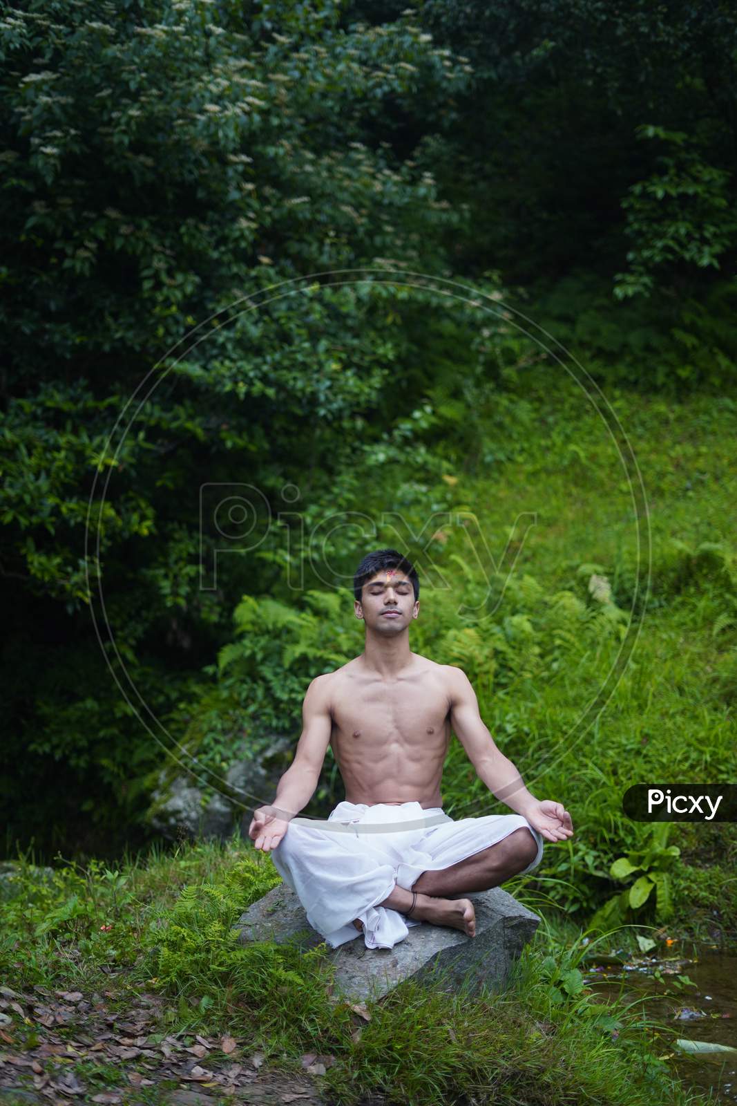 Young Indian Man Doing Yoga While Sitting On A Rock, Wearing A White Dhoti.