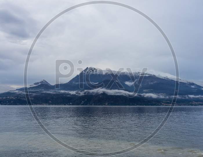 Italy, Varenna, Lake Como, A Body Of Water With A Snowcap Mountain In The Background
