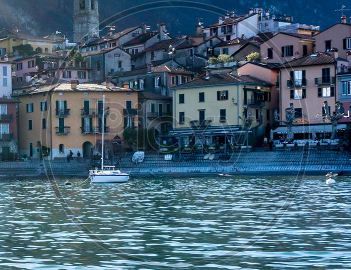 Italy, Bellagio, Lake Como, A Small Boat In A Body Of Water With Buildings In The Background
