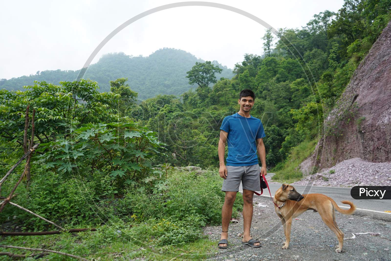 Young Man With His Dog Standing Beside A Road With Blurred Mountains In The Background.