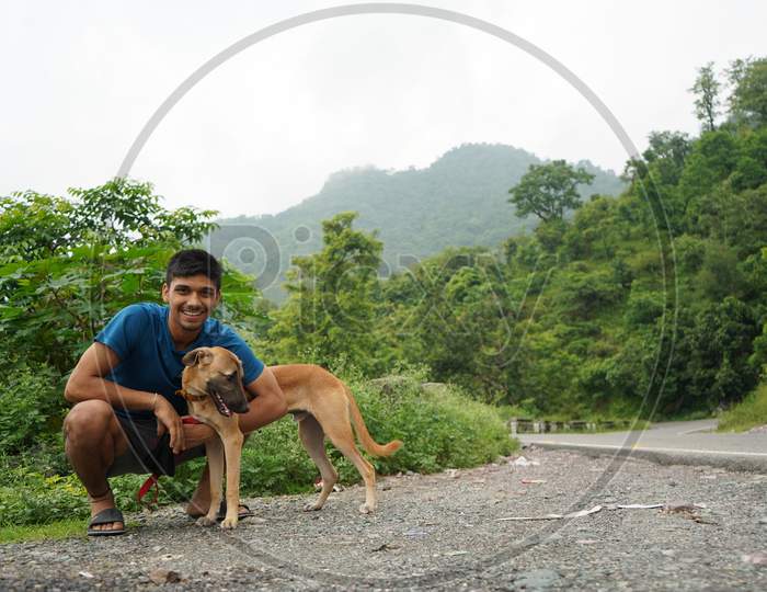 Young Man Sitting With His Dog Beside A Road With Blurred Mountains In The Background.