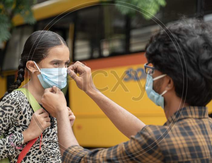 Father Helping Daughter To Wear Mask Before Getting Inside The School Bus As Coronavirus Or Covid-19 Safety Measures - Concept Of Back To School And New Normal Lifestyle.