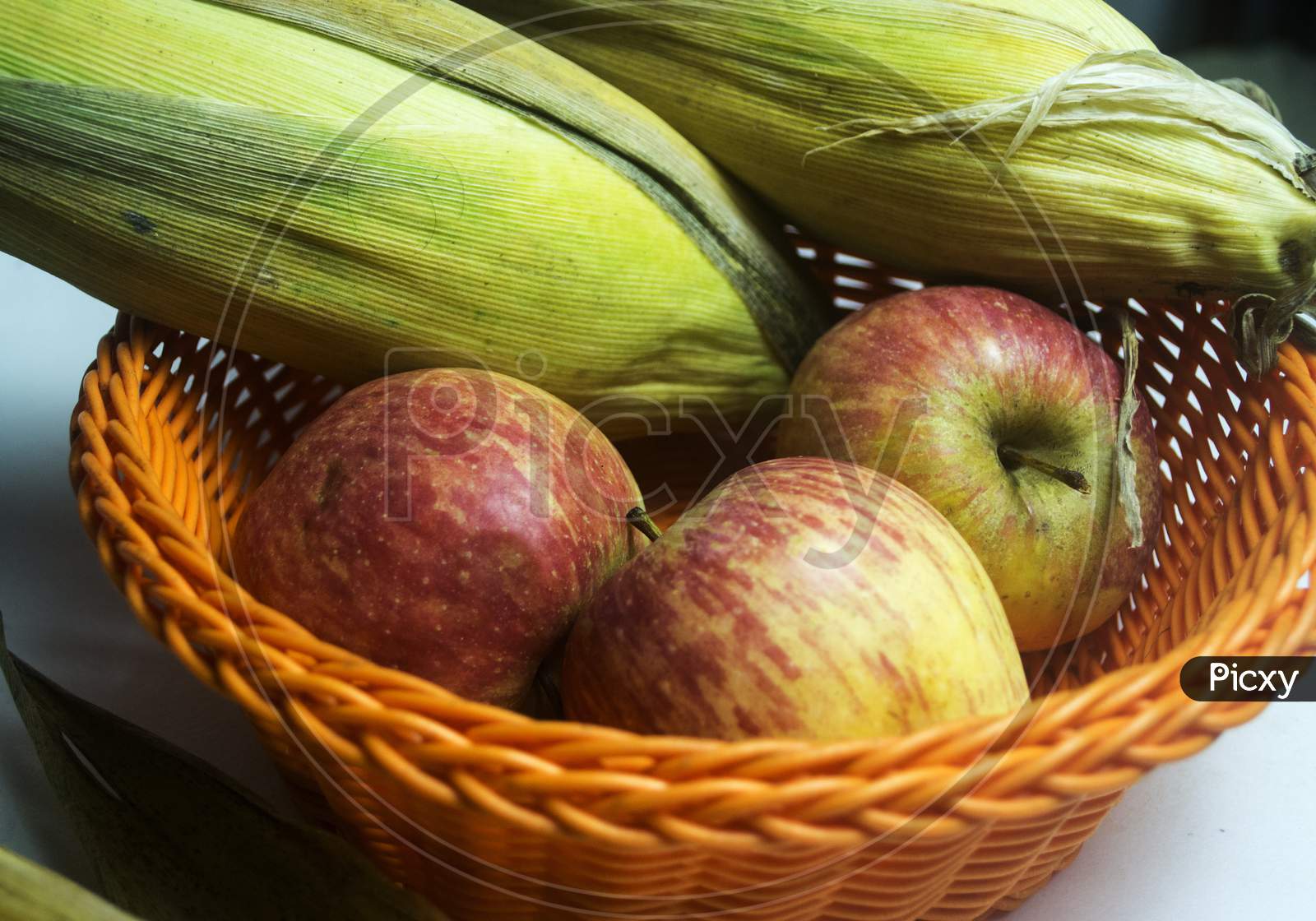 Apple And Leafed Corn In Orange Plastic Fruit Basket