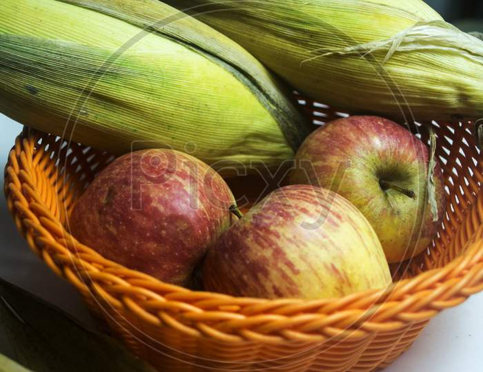 Apple And Leafed Corn In Orange Plastic Fruit Basket