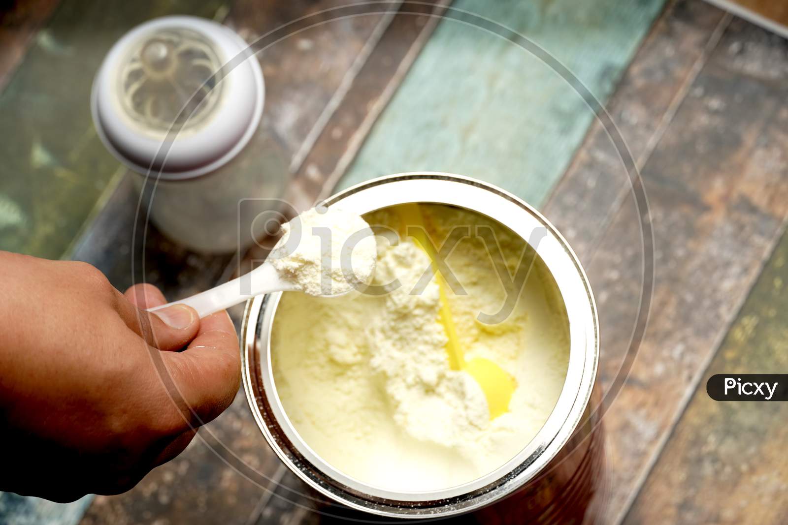 Man'S Hand Grabbing A Scoop Of Powdered Baby Milk Next To A Bottle On A Colorful Wooden Background.