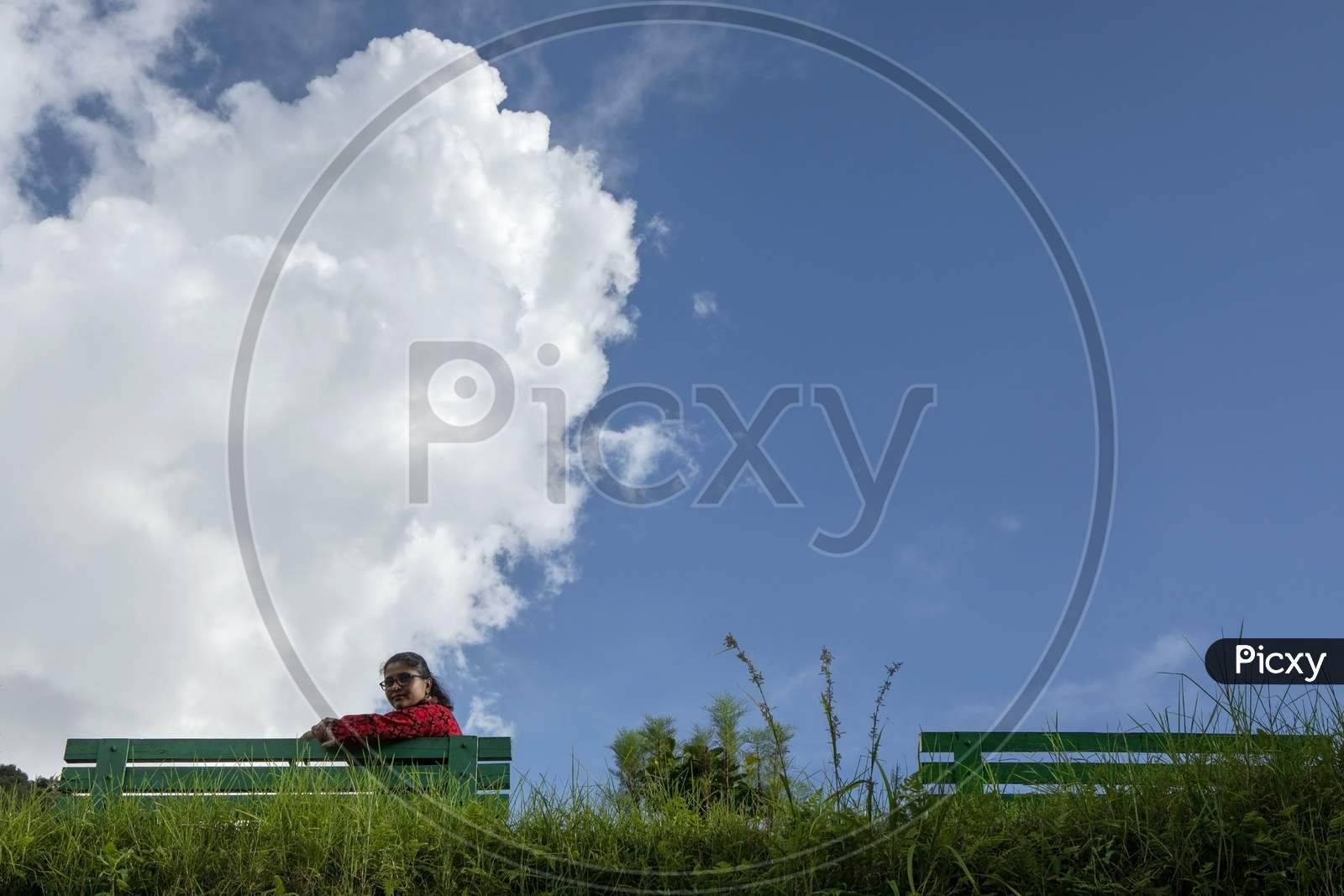 A Beautiful Young Lady In Red Dress Seating On A Green Bench In A Mountain Park In A Sunny Morning