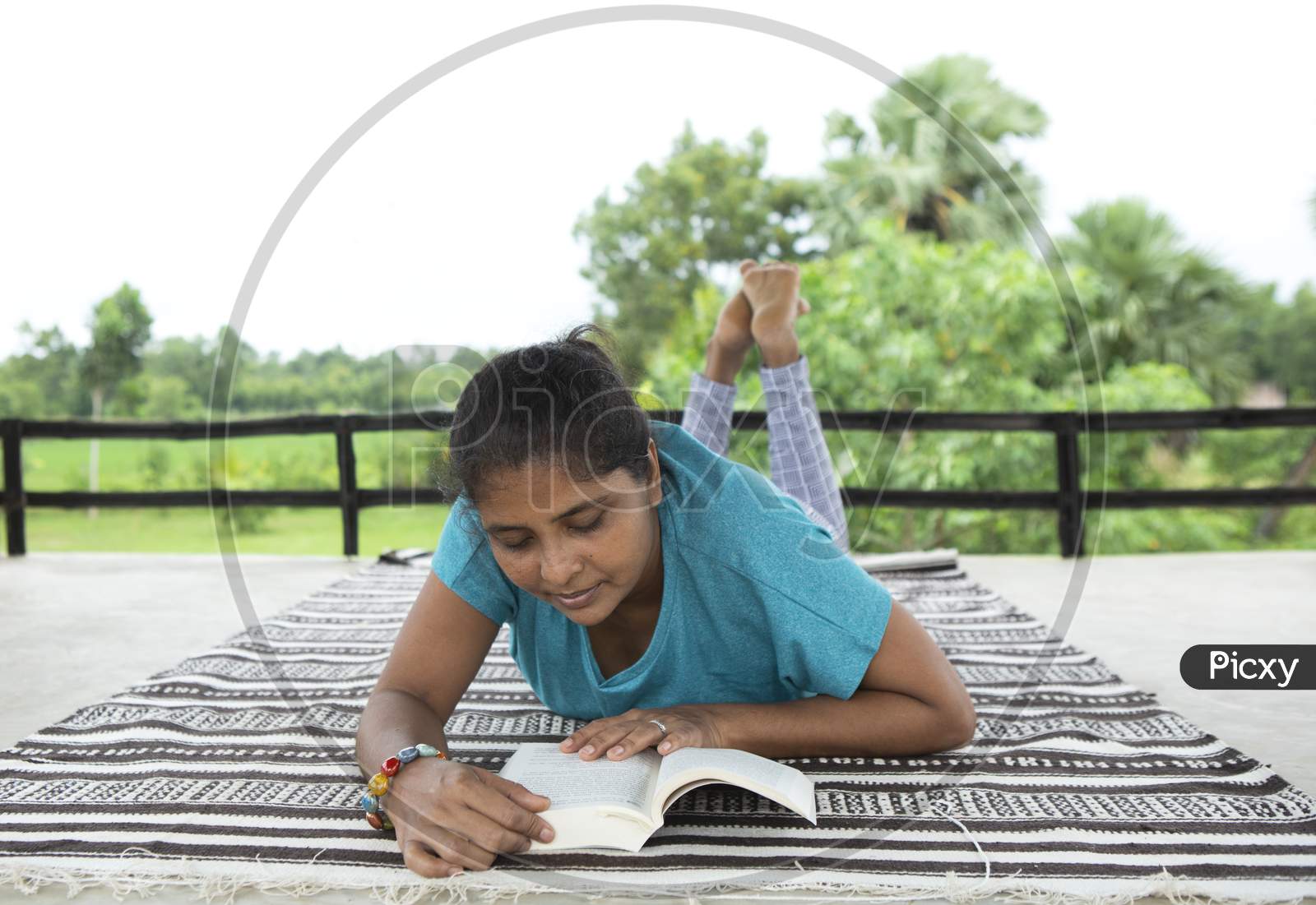 A Lady In Blue T-Shirt Reads A Book At A Peaceful Location On A Striped Mattress Surrounded By Greenery