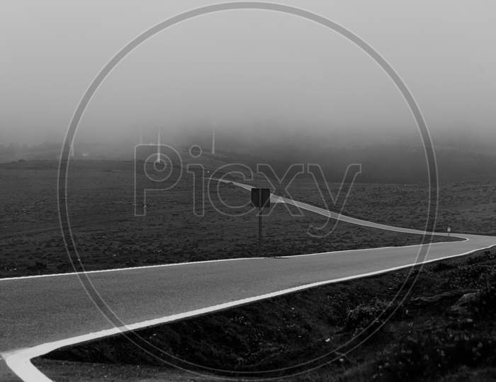 Dramatic Black And White Shot Of A Solitary Road Covered By The Mist In The Mountains With Windmills At The End Of It