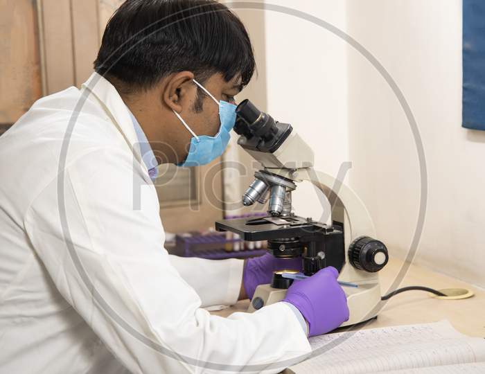Asian Indian Male Medical Doctor Or Researcher Wearing Mask Looking Through A Microscope In A Laboratory.