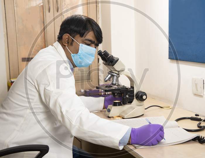Asian Indian Male Medical Doctor Or Researcher Wearing Mask Looking Through A Microscope In A Laboratory. Man Taking Note Or Doing Some Calculation At His Workplace.