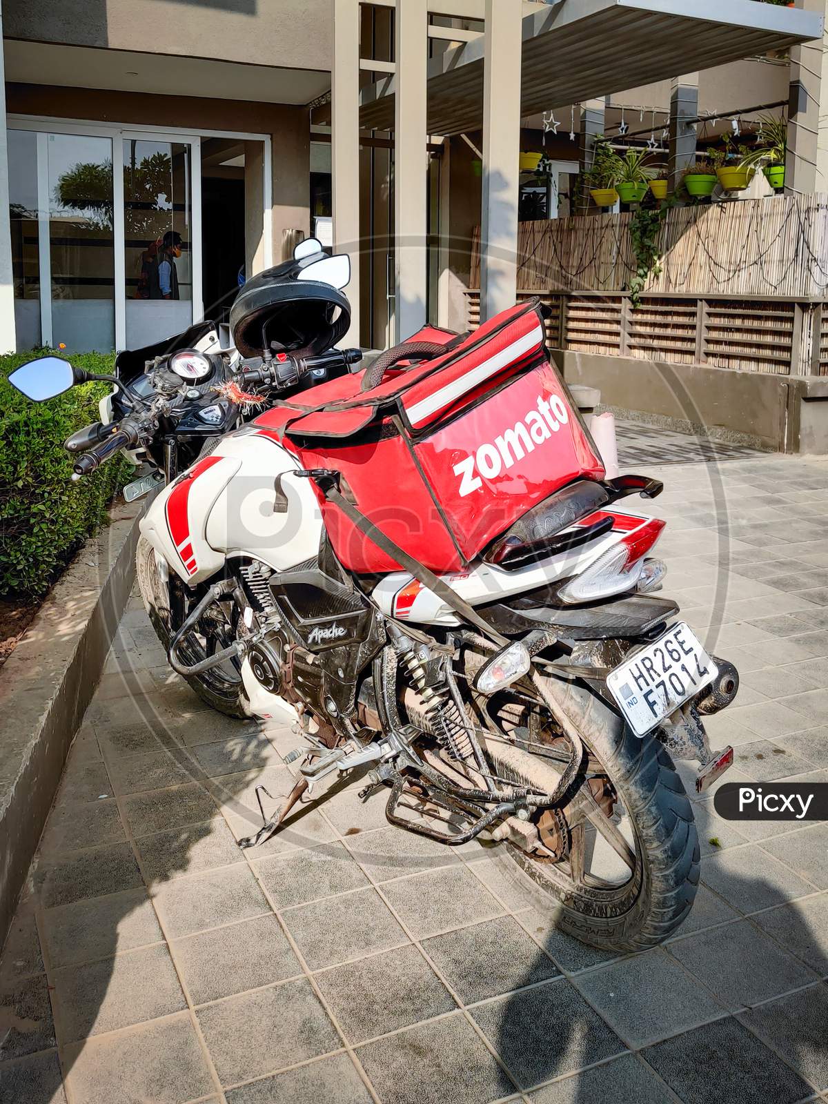 Motorcycle Rider With Red Zomato Bag Delivering Food To A Urban Complex For The Fast Growing Food Tech Startup Unicorn