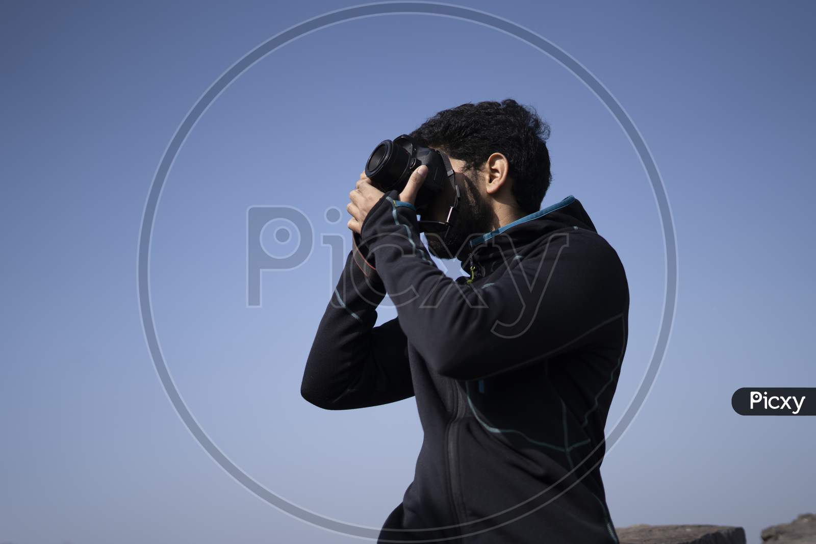 Young Indian Boy Clicking Photos From His Dslr Camera With Blue Sky In The Background.