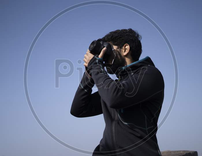 Young Indian Boy Clicking Photos From His Dslr Camera With Blue Sky In The Background.